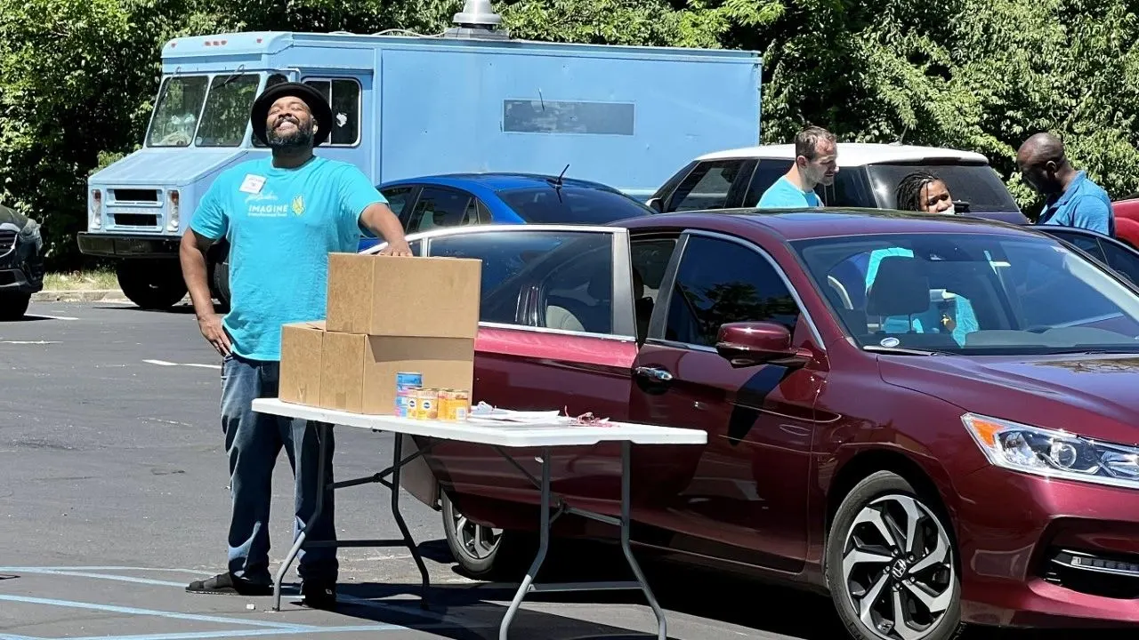 A volunteer beside boxes on a folding table as cars queue for a drive-through food distribution